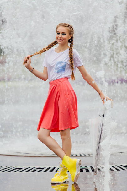 Young pretty girl with two braids in yellow boots and with transparent umbrella stands near fountain. Rainy day in city. - Photo, image