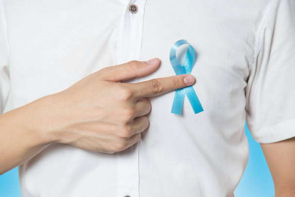 Men's healthcare concept - close up of male hand pointing to light blue ribbon for prostate cancer at left chest on blue background. - Fotografie, Obrázek