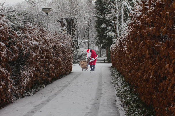 Père Noël avec sac avec des cadeaux sur le traîneau debout dans la rue dans la ville enneigée
. - Photo, image