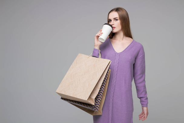 Young model long-haired blond girl in blue purple lilac jamper holds shopping paper bags and  plastic coffee glass drinking - Fotó, kép