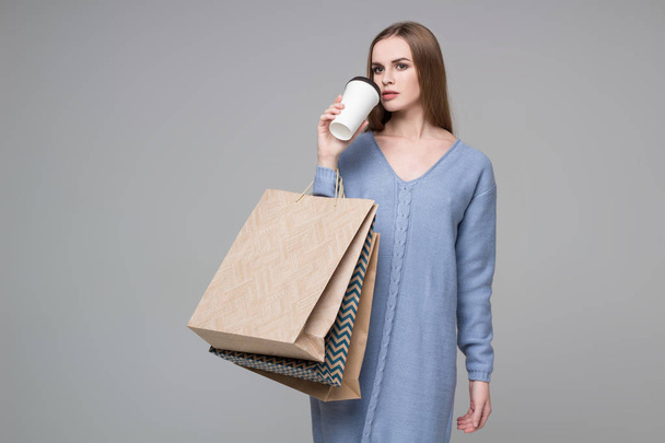 Молодая модель длинноволосая блондинка в голубом джампере держит пакеты для покупок и пластиковые кофейные стаканы
 - Фото, изображение