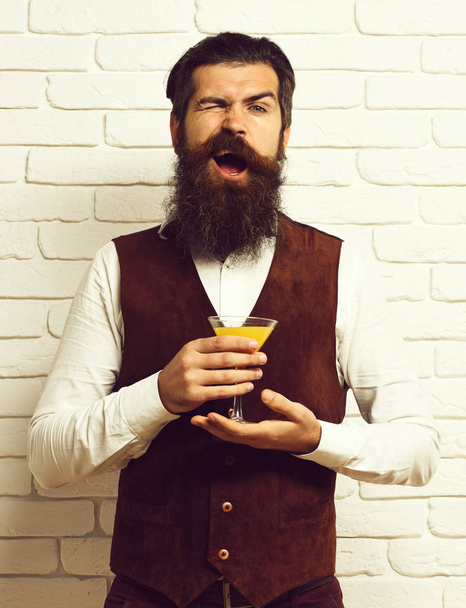 handsome bearded man with long beard and mustache has stylish hair on funny face holding glass of alcoholic beverage in vintage suede leather waistcoat on white brick wall studio background - Foto, immagini