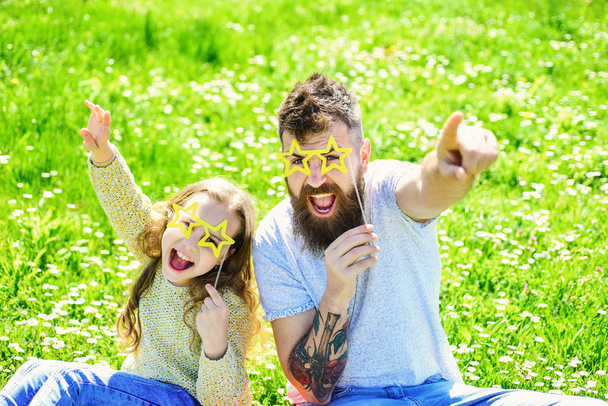 Rock star concept. Father and daughter sits on grass at grassplot, green background. Family spend leisure outdoors. Child and dad posing with star shaped eyeglases photo booth attribute at meadow - Photo, image