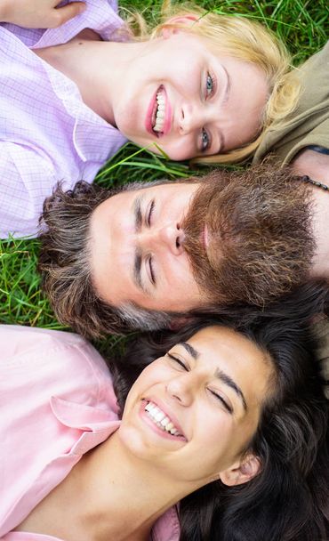Man fall in love both women. Happy threesome. Hipster relaxing pleasant women company. Blonde and brunette lay on his shoulders. Man bearded hipster lay on grass with two girls blonde and brunette - Foto, Imagem