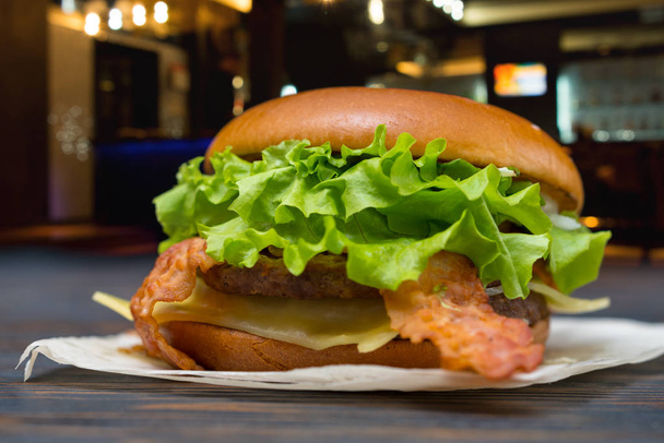 Sabroso queso y hamburguesa de tocino con lechuga fresca recortando en una vista de ángulo bajo en una servilleta de papel al aire libre en una mesa en la calle de la ciudad por la noche
 - Foto, Imagen