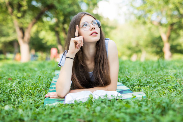 Concentrated young woman resting with book in park. Serious beautiful girl lying on grass while reading favorite romance. - Foto, Imagem
