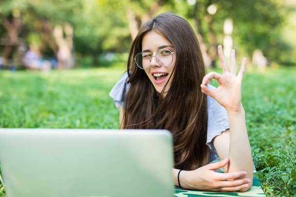 Happy brunette woman in eyeglasses lying on grass in park with laptop computer and showing ok sign - 写真・画像