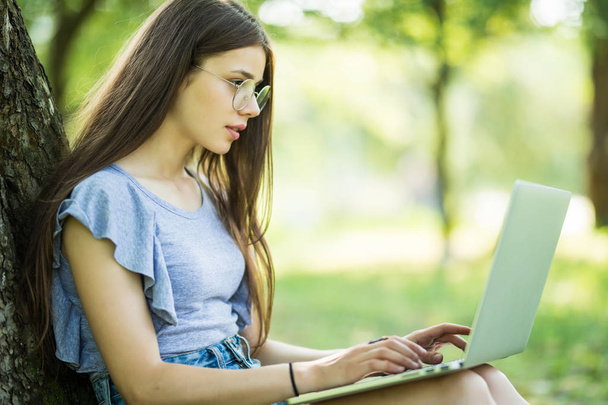 Portrait of young woman sitting on green grass in park with legs crossed during summer day while using laptop - Photo, Image