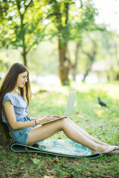 Young beautiful woman is sitting on green grass under the tree in the garden and working on her laptop - Zdjęcie, obraz