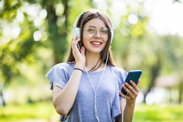 S úsměvem mladá studentka s batohem držení mobilního telefonu, procházky v parku, poslechu hudby pomocí sluchátek - Fotografie, Obrázek