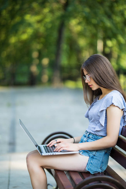 Side view of pleased brunette woman in eyeglasses sitting on bench in park and using laptop computer - Foto, Imagen