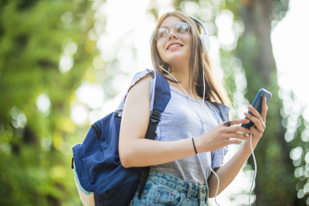 Smiling attractive young woman with backpack listening to music from cell phone in park - 写真・画像