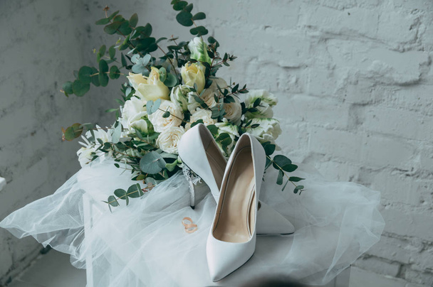 Γαμήλια παπούτσια νύφη ντυμένη στα λευκά, με ένα μπουκέτο από λευκά τριαντάφυλλα και άλλα λουλούδια, γαμήλια δαχτυλίδια σε ένα σκαμνί - Φωτογραφία, εικόνα
