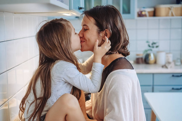 Mom kisses her little daughter in the kitchen - Фото, изображение