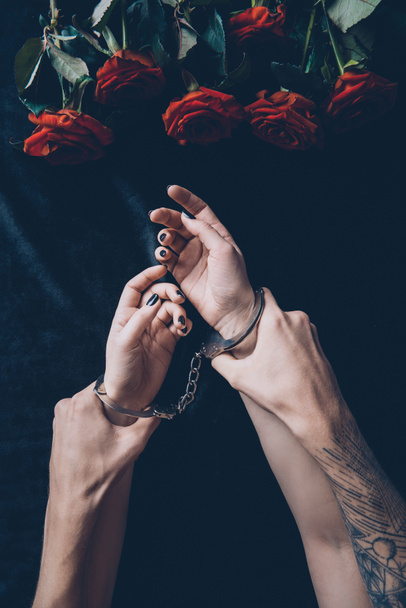 cropped shot of man holding female hands in handcuffs above black fabric with red roses - Photo, Image