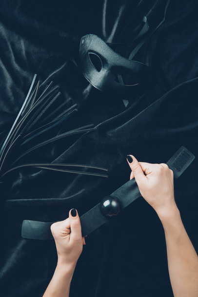Schnappschuss weiblicher Hände mit Knebel, Lederpeitsche und Maske auf schwarzem Stoff - Foto, Bild