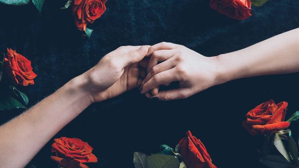 bijgesneden schot van paar hand in hand boven zwarte stof met rode rozen - Foto, afbeelding