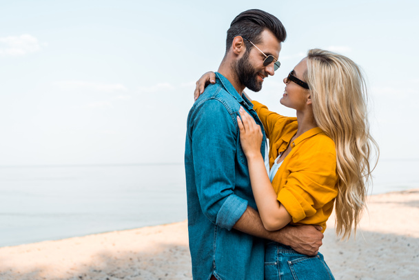 side view of couple in sunglasses hugging and looking at each other near ocean - Фото, зображення