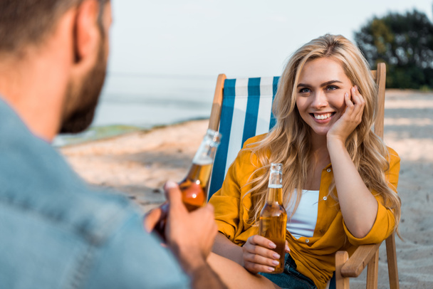 para siedzi na leżakach z piwa w butelkach na piaszczystej plaży, uśmiechnięta dziewczyna patrząc na chłopaka - Zdjęcie, obraz
