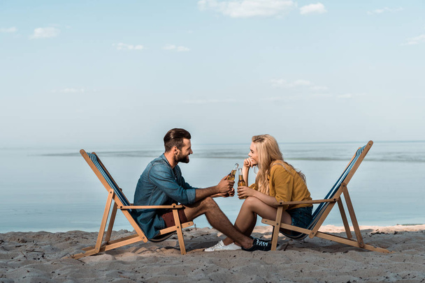 vista lateral de la novia y el novio sentado en tumbonas y tintineo con botellas de vidrio de cerveza en la playa de arena
 - Foto, Imagen