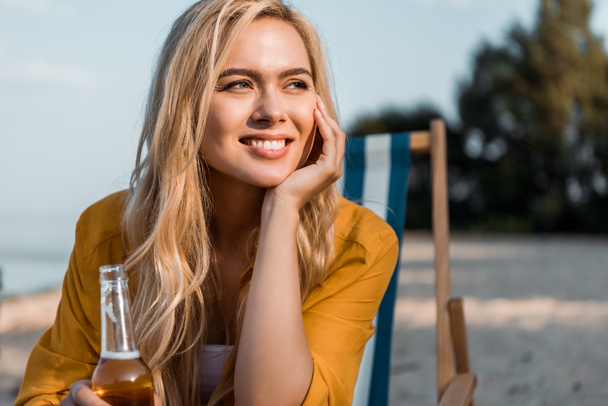 atractiva chica sonriente sentada en la tumbona con cerveza en botella y mirando hacia otro lado
 - Foto, imagen