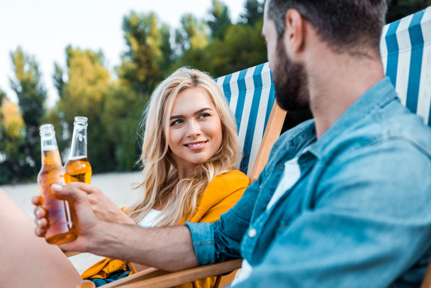 couple sitting on sun loungers and clinking with glass bottles of beer on sandy beach - Photo, Image
