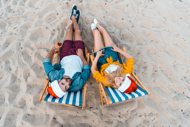 vista de ángulo alto de pareja en sombreros de santa mentira en tumbonas con cerveza en botellas en la playa de arena
 - Foto, Imagen