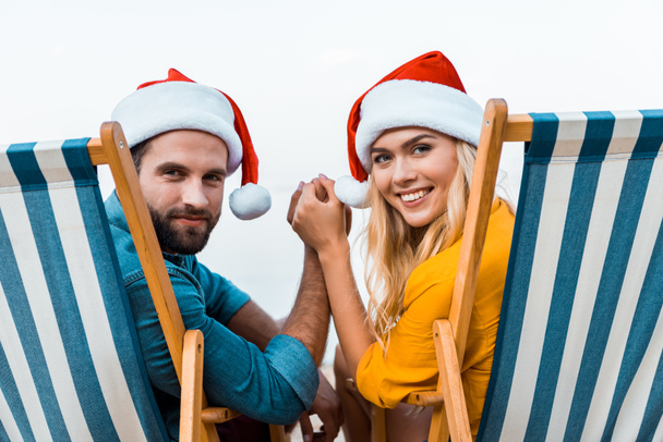 Portret van paar in santa hoeden zittend op de ligstoelen, houdend handen en kijken naar camera op strand - Foto, afbeelding
