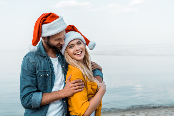 smiling boyfriend in santa hat hugging girlfriend on beach - Fotoğraf, Görsel