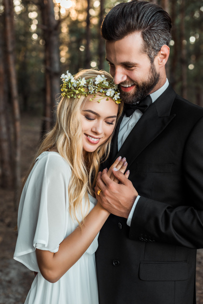 schöne glückliche Braut im weißen Kleid und schöner Bräutigam im Anzug umarmen und Händchen halten im Wald  - Foto, Bild