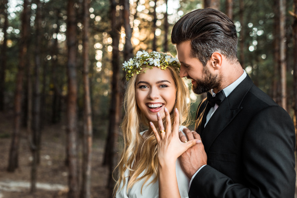 happy bride in white dress and wreath showing wedding ring on finger in forest - Photo, Image