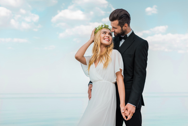 ніжна весільна пара в костюмі і білій сукні тримає руки і дивиться один на одного на пляжі
 - Фото, зображення