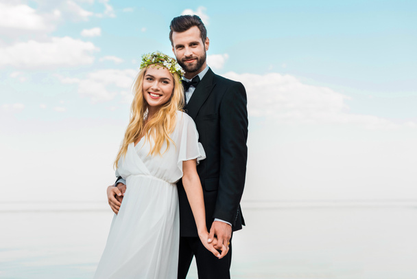 wedding couple in suit and white dress holding hands and looking at camera on beach - Photo, Image