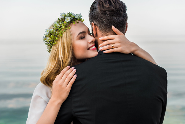 attractive bride with closed eyes and groom in suit hugging on beach - Φωτογραφία, εικόνα