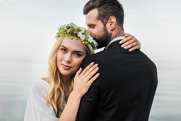 attractive bride in wreath and groom in suit cuddling on beach - Photo, Image