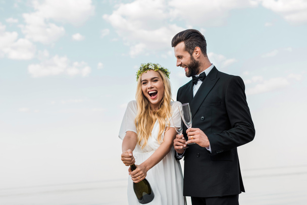 happy bride in white dress opening champagne bottle on beach - Фото, зображення