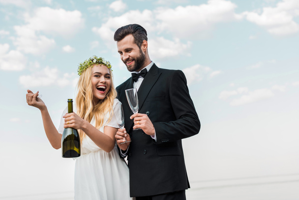 laughing bride in white dress opening champagne bottle on beach - Foto, Bild