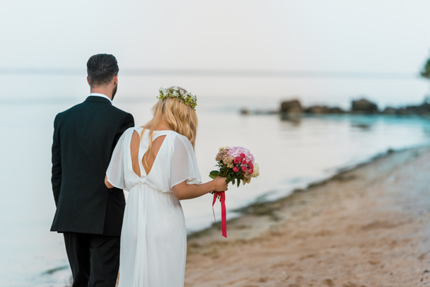 Achteraanzicht van bruidspaar met boeket wandelen op strand - Foto, afbeelding