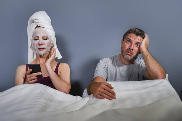 funny lifestyle portrait of eccentric and weird housewife with makeup facial mask and towel using mobile phone in bed and husband in desperate face expression in man woman relationship concept  - Foto, Imagen