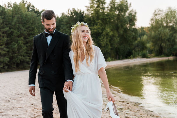 手を繋いでいるカップルを結婚式や花嫁持株ハイヒールを手に笑い、ビーチの上を歩く - 写真・画像