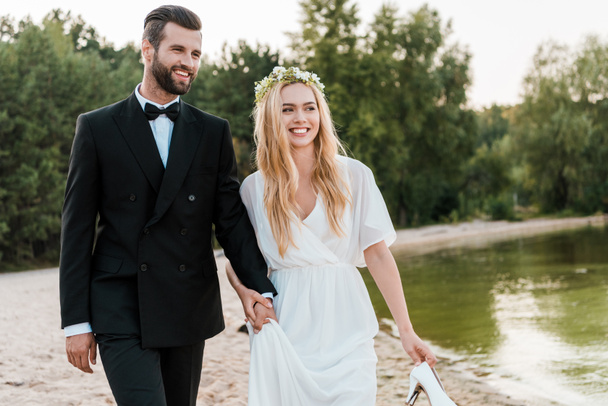 smiling wedding couple holding hands and walking on beach, bride holding high heels in hand - Zdjęcie, obraz