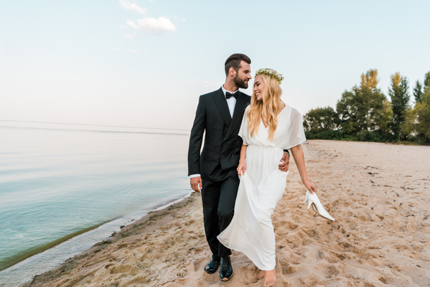 Hochzeitspaar umarmt und geht am Sandstrand spazieren, Braut hält High Heels in der Hand - Foto, Bild
