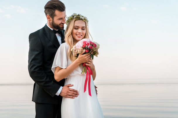 Bräutigam umarmt Braut und sie hält Brautstrauß am Strand - Foto, Bild