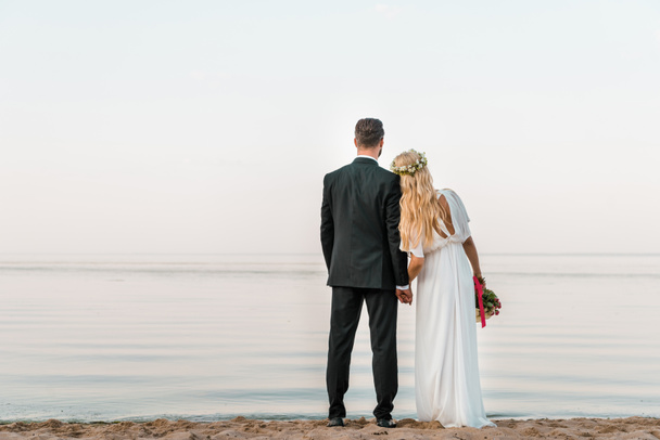 結婚式のカップルの結婚式のブーケとビーチの上に立って、海を見ての背面図 - 写真・画像
