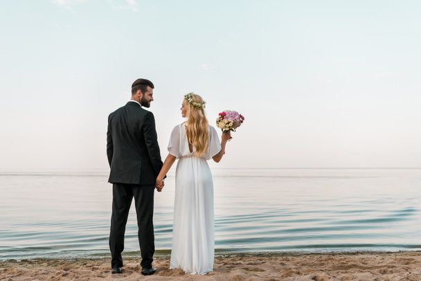 Rückansicht des Hochzeitspaares, das mit Brautstrauß am Strand steht und einander ansieht - Foto, Bild