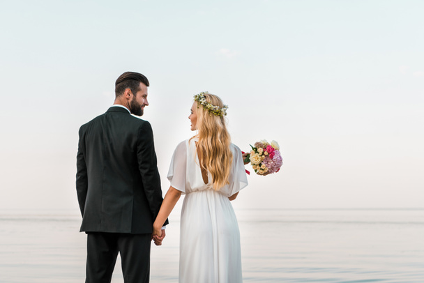 vista trasera de la pareja de boda de pie en la playa, tomados de la mano y mirando el uno al otro
 - Foto, imagen