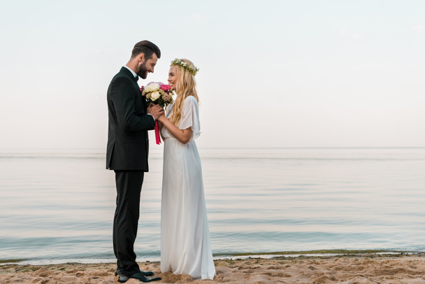vista lateral do casal de casamento afetuoso de pé com buquê na praia, noiva farejando rosas
 - Foto, Imagem