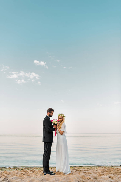 side view of wedding couple standing with bouquet on sandy beach, bride sniffing roses - Photo, Image
