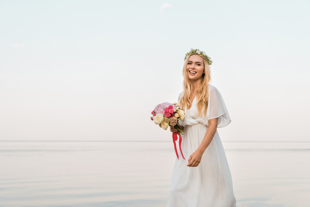 smiling attractive bride in white dress and wreath holding wedding bouquet and walking near sea - Fotó, kép