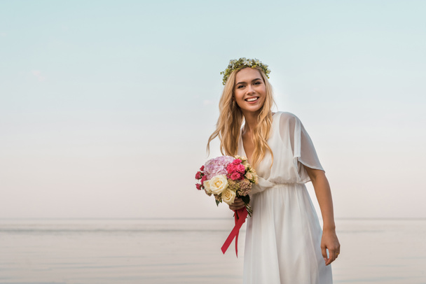 smiling attractive bride in white dress and wreath holding wedding bouquet of roses and looking at camera on beach - Photo, Image